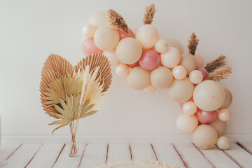 A delicate photo zone with an arch of pink and beige balls is decorated with decorative leaves....