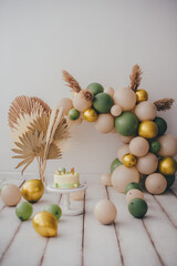 Stylish photo zone with a cake and an arch of beige, green and gold balloons. Light background white wood floor.