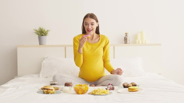 Pregnancy food caprices. Young excited pregnant lady sitting on bed and enjoying sweet junk donut, slow motion