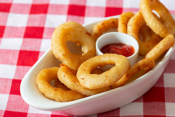 Battered onion rings in a dish,  with a tomato ketchup dip.  On a gingham tablecloth