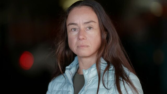 In front, a close-up portrait of an adult European woman with long hair standing on the street and looking amiably into the camera. Human face without makeup