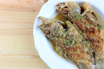 Thai Giant gourami fish and garlics deep fried and sweet spicy sauce in white dish on pine wooden table. Deep fried fish with Sweet chili. Thai food fried fish with spicy sauce. Thai style food.