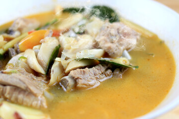 Spicy boiled Pork bone and straw mushroom, wild tomato, herb in tom yum soup on white bowl on wooden table. Tom Yum soup pork in Thai style food.