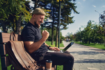 Obraz premium happy victorious bearded man sit on the bench outdoor while look at laptop gesturing yes win winner 