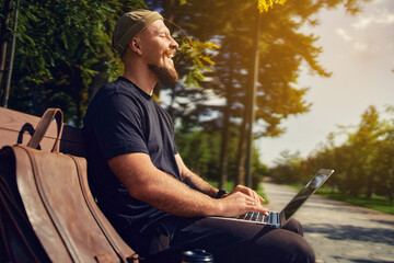 Outdoor photo of laughing millennial in glasses is sitting on a bench while working at a laptop pc 