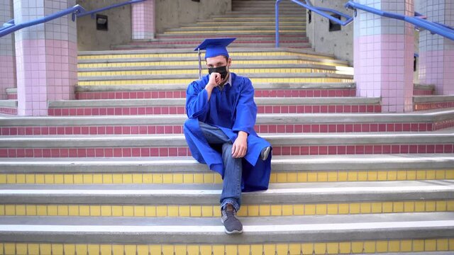 Male graduate sits at school in his cap and gown with face-mask on