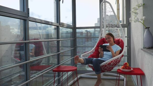 Barefoot pretty woman in blue t-shirt and jeans puts tablet on red coffee table, gets up from wicker hanging chair and leaves. Cozy place relax on home or hotel loggia with panoramic windows.