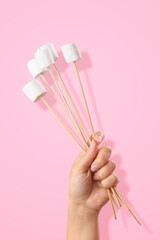 A female hand holds a group of skewers with marshmallows on a pink background.
