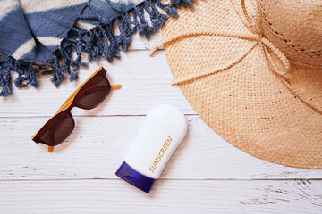 top view of brown floppy straw hat for lady with blue tie dye beach shawl ,sunglasses and sunscreen bottle on white wooden background , summer and holiday concept
