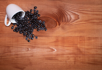 A white coffee cup lies and coffee beans pour out of it on the texture of natural wood: a place for text, a morning invigorating drink for coffee shops