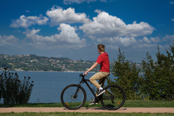 Obraz premium The movement of a cyclist in a red T-shirt along the coastline in the background lake, mountains, blue sky with clouds. A man on a mountain bike lake garda.