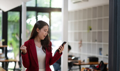 Smiling Asian woman holding mobile phone with fist hand and excited for success in office