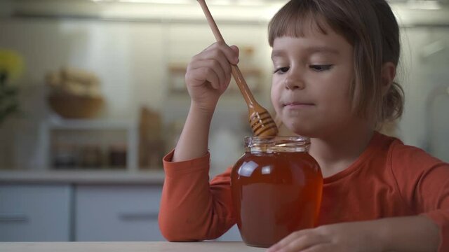 ittle child girl eat honey as it drops on her tongue licking it out from the wooden spoon. Kid eats honey from a jar at home in the kitchen. Healthy organic sweets for kids