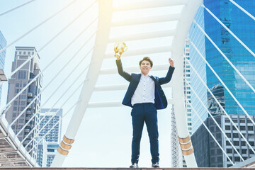 Successful businessman celebrating his victory with arms up and holding gold trophy on cityscape background.