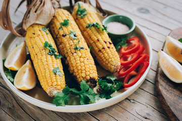 Baked corn cobs with sauce, coriander, sliced bell pepper, lemon and salt on wooden table. Mexican food. Top view