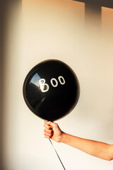 Girl's hand holding black baloon with word Boo on white wall background. Halloween concept