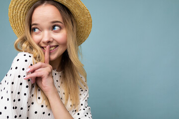 Closeup photo of young positive happy cute beautiful blonde woman with sincere emotions wearing summer dress and straw hat isolated over blue background with copy space and showing silence gesture