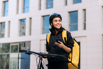 Food delivery woman using smart phone on street in city. Female courier in cycling helmet with backpack looking away. - obrazy, fototapety, plakaty
