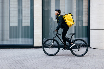 Side view of a young woman riding her bike in the city. Courier with a thermal backpack in her way...