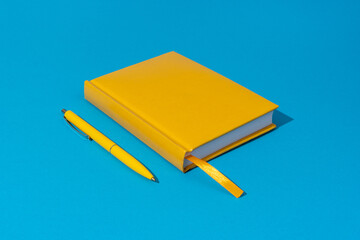 Photo of closed yellow notebook and ball-point pen over blue background. Minimalist image of closed...