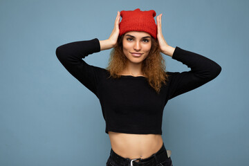 Photo of young pretty positive brunette woman curly with sincere emotions wearing stylish black crop top and red hat isolated on blue background with copy space