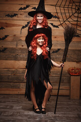 Fototapeta na wymiar Halloween Concept - Beautiful caucasian mother and her daughter with long red hair in witch costumes celebrating Halloween posing with over bats and spider web on Wooden studio background.