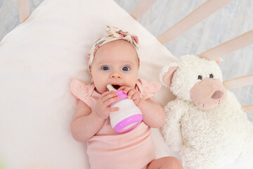 baby girl sucks a bottle of milk in the crib lying on her back with a toy in the nursery, baby food concept