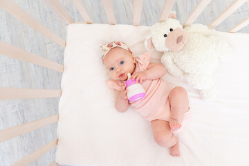 baby girl sucks a bottle of milk in the crib lying on her back with a toy in the nursery, baby food...
