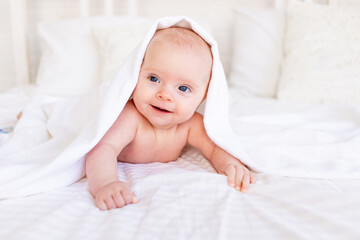 Fototapeta na wymiar baby girl in a towel after bathing in a crib on a white cotton bed lying on her stomach smiling in the nursery, newborn hygiene