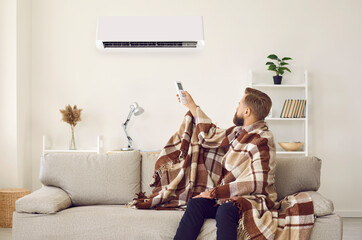 Sad angry man sitting on sofa trying to warm up under plaid while freezing in cold living room with...
