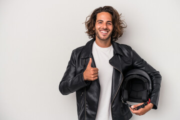Young biker caucasian man holding a motorbike helmet isolated on gray background smiling and...