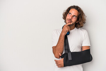 Young caucasian man with broken hand isolated on gray background looking sideways with doubtful and...