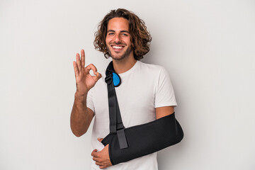 Young caucasian man with broken hand isolated on gray background cheerful and confident showing ok...