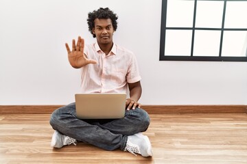 African man with curly hair using laptop sitting on the floor doing stop sing with palm of the hand. warning expression with negative and serious gesture on the face.