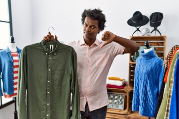 African man with curly hair holding shirt from clothing rack at retail shop with angry face, negative sign showing dislike with thumbs down, rejection concept