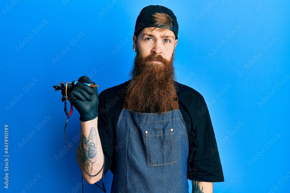 Sticker Redhead man with long beard tattoo artist wearing professional uniform and gloves relaxed with serious expression on face. simple and natural looking at the camera.