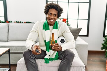 Young african american hooligan man drinking beer supporting soccer team sitting on the sofa at home.