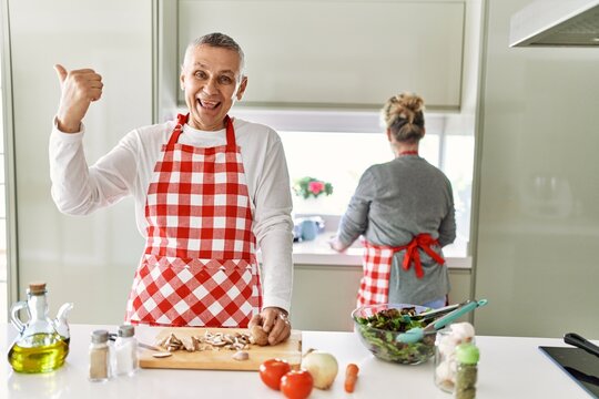 Middle age caucasian couple cooking healthy salad pointing thumb up to the side smiling happy with open mouth