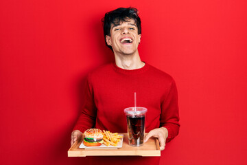 Handsome hipster young man eating a tasty classic burger with fries and soda celebrating crazy and amazed for success with open eyes screaming excited.