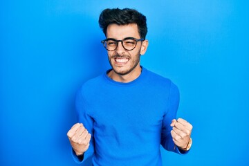 Naklejka premium Young hispanic man wearing casual clothes and glasses very happy and excited doing winner gesture with arms raised, smiling and screaming for success. celebration concept.