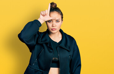 Young hispanic girl wearing sportswear making fun of people with fingers on forehead doing loser...