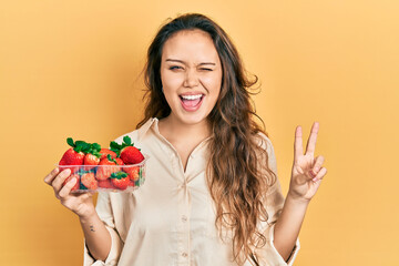 Young hispanic girl holding strawberries smiling with happy face winking at the camera doing...