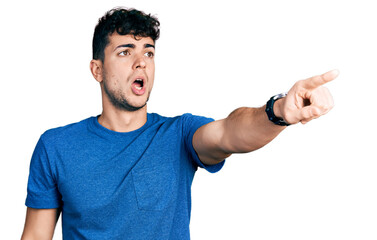 Young hispanic man wearing casual t shirt pointing with finger surprised ahead, open mouth amazed expression, something on the front