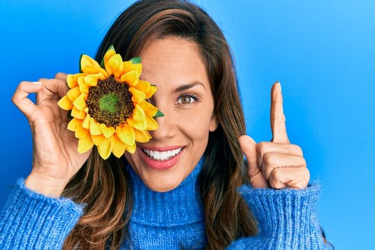 Young latin woman holding sunflower over eye smiling with an idea or question pointing finger with happy face, number one
