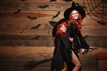 Fototapeta na wymiar Halloween Concept - Beautiful caucasian mother and her daughter with long red hair in witch costumes flying with magic broomstick.