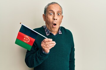 Handsome senior man with grey hair holding afghanistan flag scared and amazed with open mouth for surprise, disbelief face