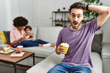 Hispanic father of interracial family drinking a cup coffee crazy and scared with hands on head, afraid and surprised of shock with open mouth