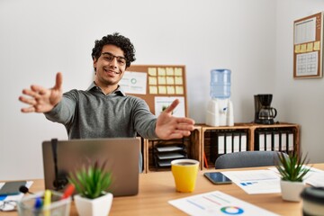 Young hispanic man wearing business style sitting on desk at office looking at the camera smiling with open arms for hug. cheerful expression embracing happiness.