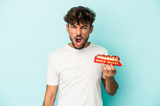 Young arab man holding a hotdog isolated on blue background screaming very angry and aggressive.