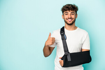 Young arab man with broken hand isolated on blue background smiling and raising thumb up